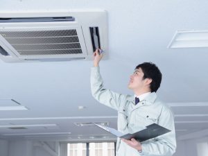 A technician in a light uniform inspects and adjusts a ceiling-mounted air conditioning unit while holding a clipboard in an office setting.