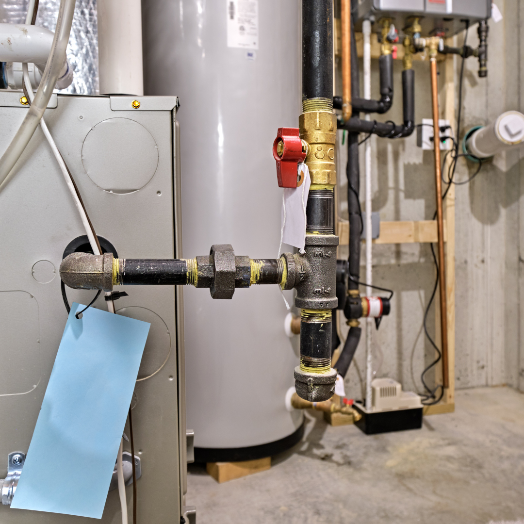 Gas and water pipes connected to a furnace and water heater in a basement utility room illustrate a recent heating installation, with a blue tag attached to one of the pipes.