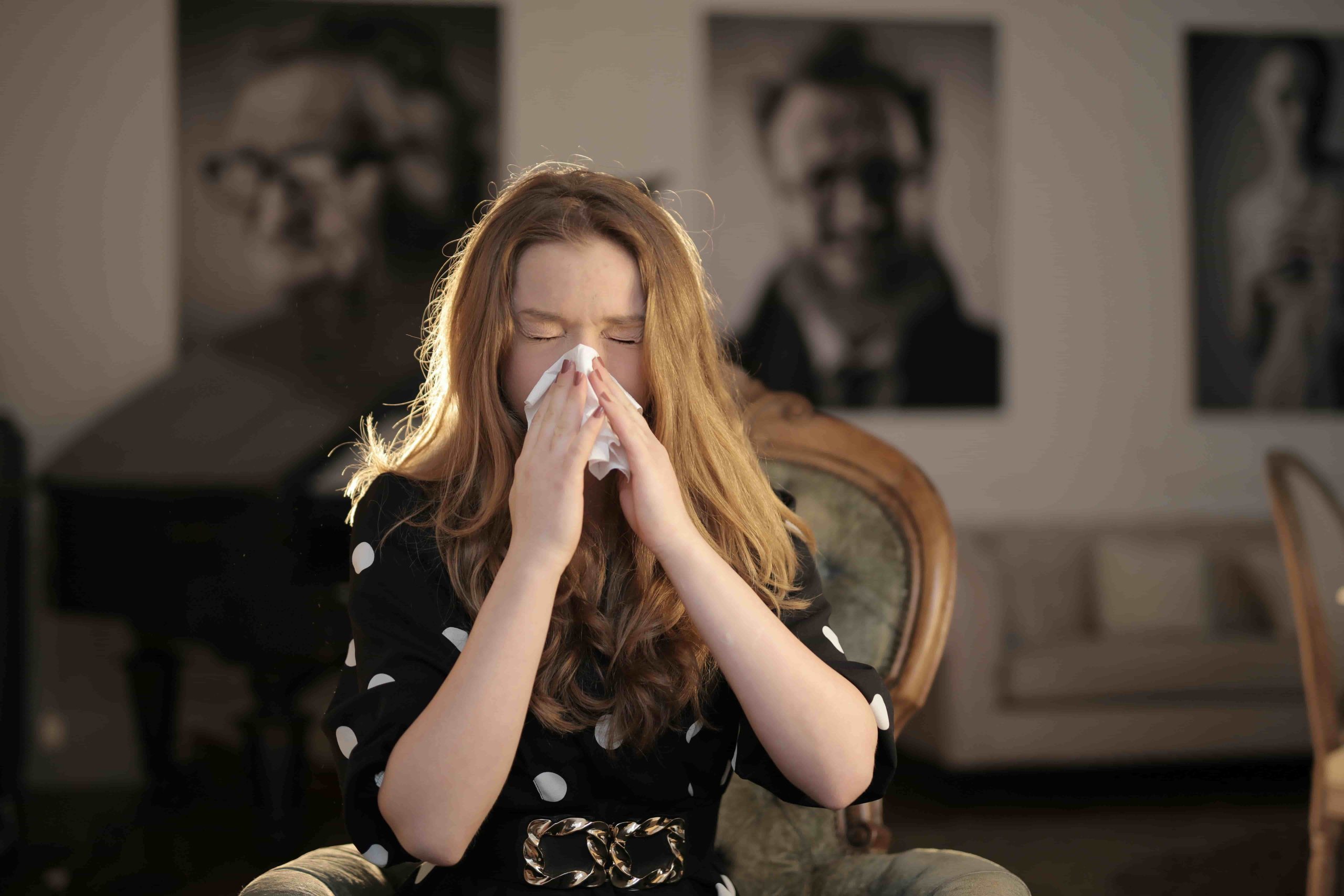 A woman with wavy hair sits on a chair indoors, holding a tissue to her nose as if sneezing or blowing her nose—perhaps searching for the Best Air Filters for Allergy Relief in Costa Mesa, with blurred portraits in the background.