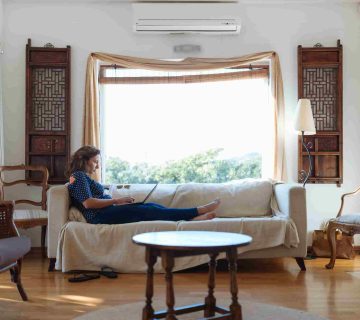 A woman sits on a white sofa using a laptop in a bright living room with wooden furniture and large windows, searching for tips on how to choose the right size AC for your Irvine home.