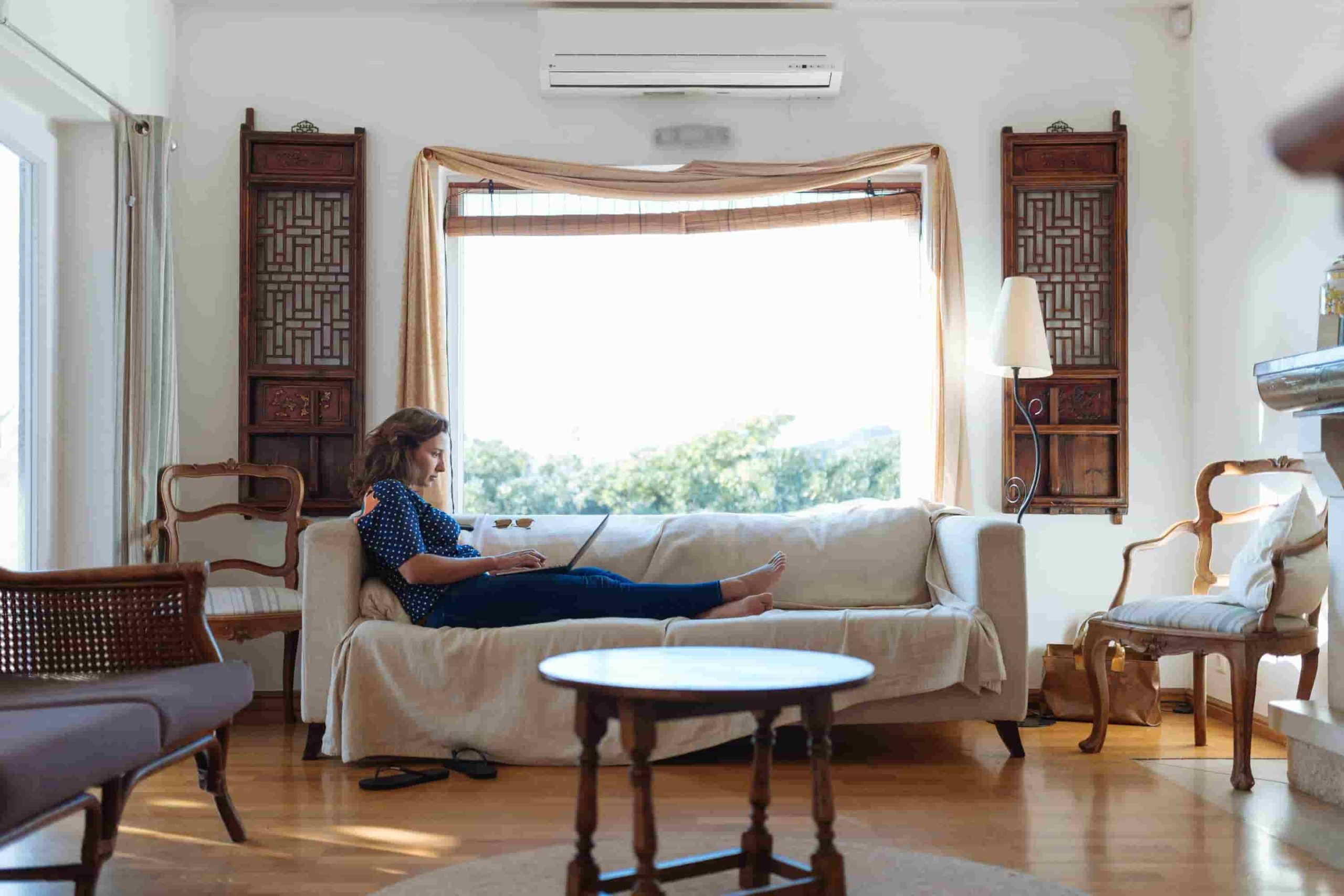 A woman sits on a white sofa using a laptop in a bright living room with wooden furniture and large windows, searching for tips on how to choose the right size AC for your Irvine home.