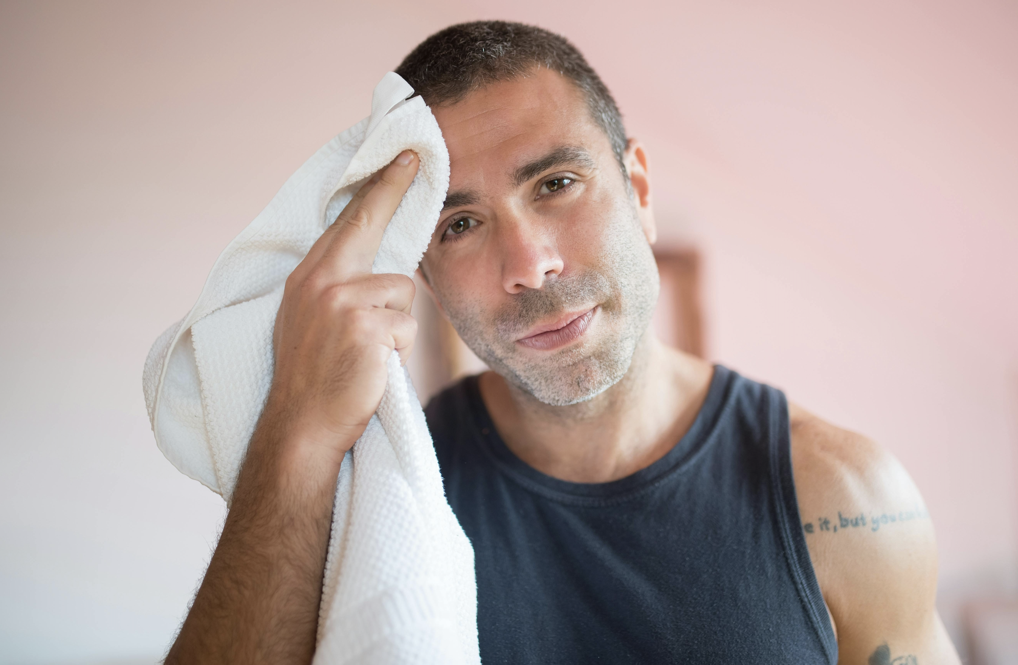 A man in a sleeveless shirt wipes sweat from his forehead with a white towel, looking directly at the camera—perhaps wondering, What Causes AC to Stop Cooling in Irvine?.