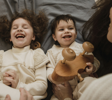 Two young children lie on a bed, smiling and laughing, while an adult sits nearby. One child holds a wooden toy—reminding us to consider what are the health risks of poor indoor air quality for children in Irvine.