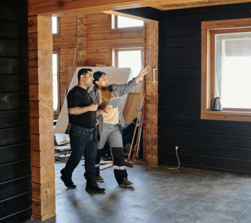 Two people stand in a partially finished wooden interior in Irvine, one gesturing toward the ceiling while discussing construction and asking, "How often should air ducts be inspected in Irvine, CA?.