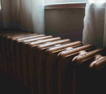 A metal radiator is positioned below a window with white curtains, illuminated by soft natural light—an inviting scene, but watch for signs your furnace needs repair in Newport Beach homes to keep spaces cozy and comfortable.