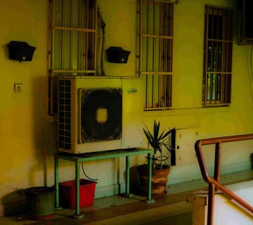 A wall mounted air conditioning unit sits on a metal stand beside potted plants, near barred windows and a handrail in an indoor space—ideal for checking signs of AC Capacitor Failure: Symptoms and Replacement Costs 