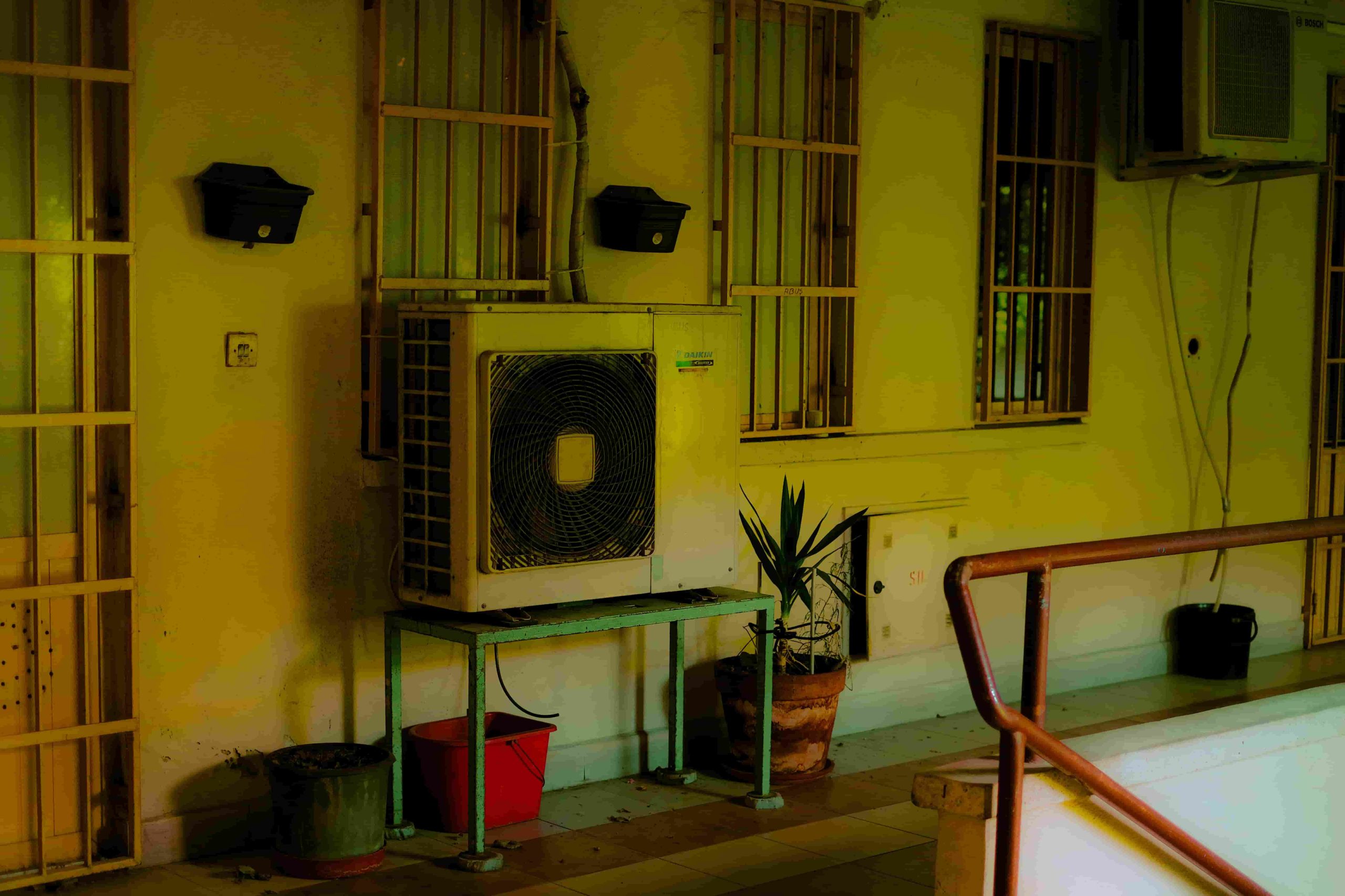 A wall mounted air conditioning unit sits on a metal stand beside potted plants, near barred windows and a handrail in an indoor space—ideal for checking signs of AC Capacitor Failure: Symptoms and Replacement Costs