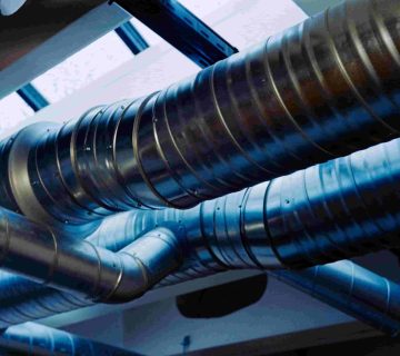 Large metal air ducts run across a ceiling near skylights, with blue tinted light reflected on their surface—an ideal spot to assess how to know if air ducts need repair or replacement 