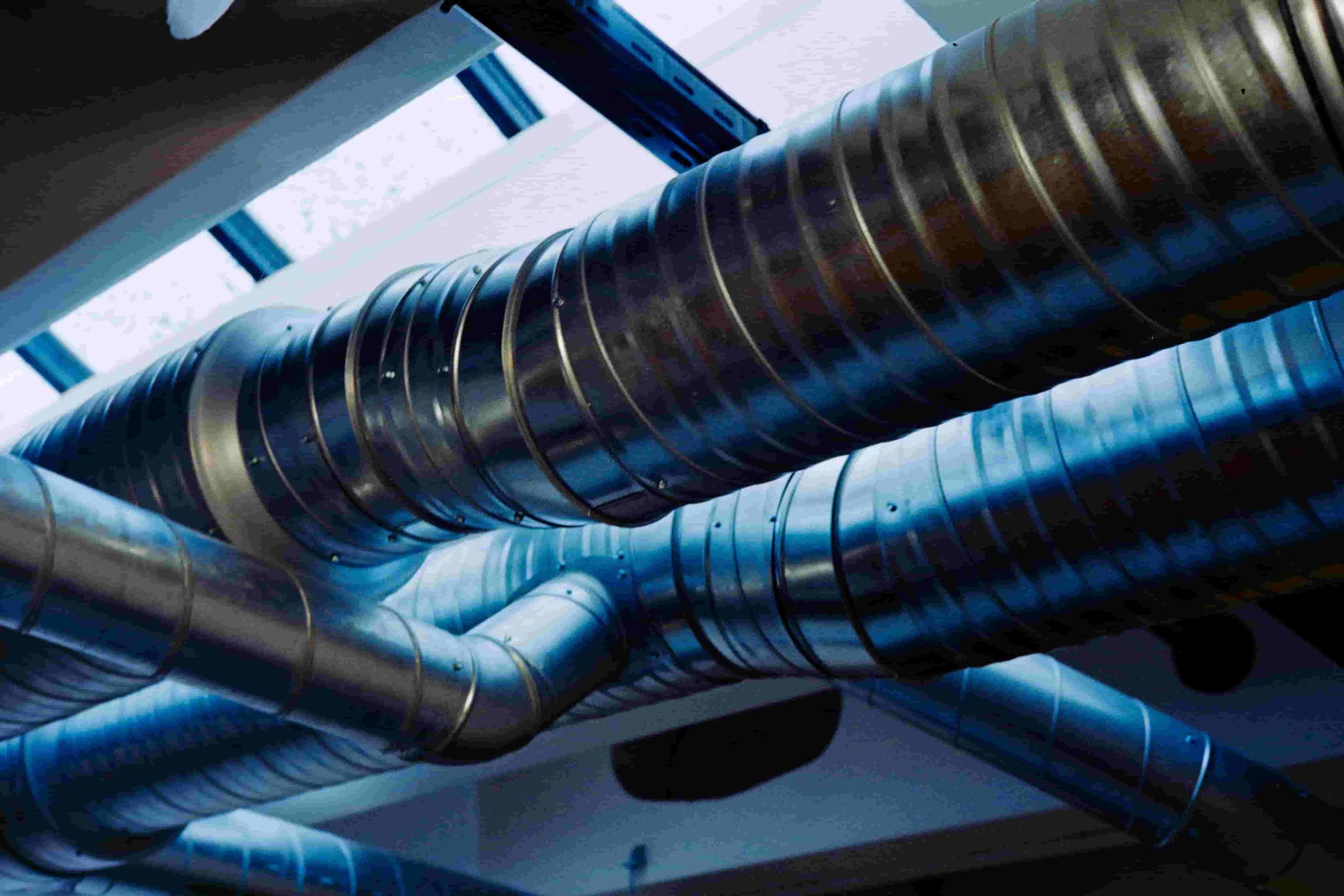 Large metal air ducts run across a ceiling near skylights, with blue tinted light reflected on their surface—an ideal spot to assess how to know if air ducts need repair or replacement 