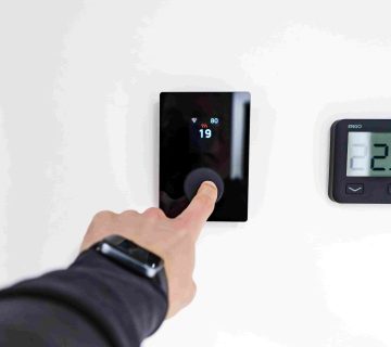 A person adjusts a digital thermostat on a white wall, demonstrating how smart thermostats can help lower summer energy bills; another display shows a temperature of 22 5°C 