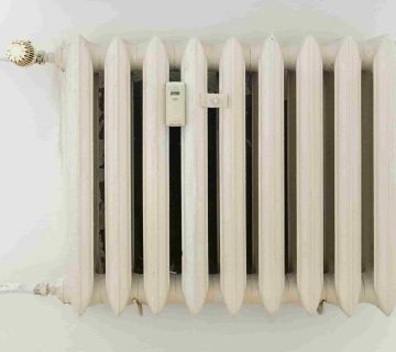 A white metal radiator with seven vertical sections, viewed from above and connected by pipes on both ends against a plain wall, is a subtle reminder of the cost of ignoring annual furnace tune ups 