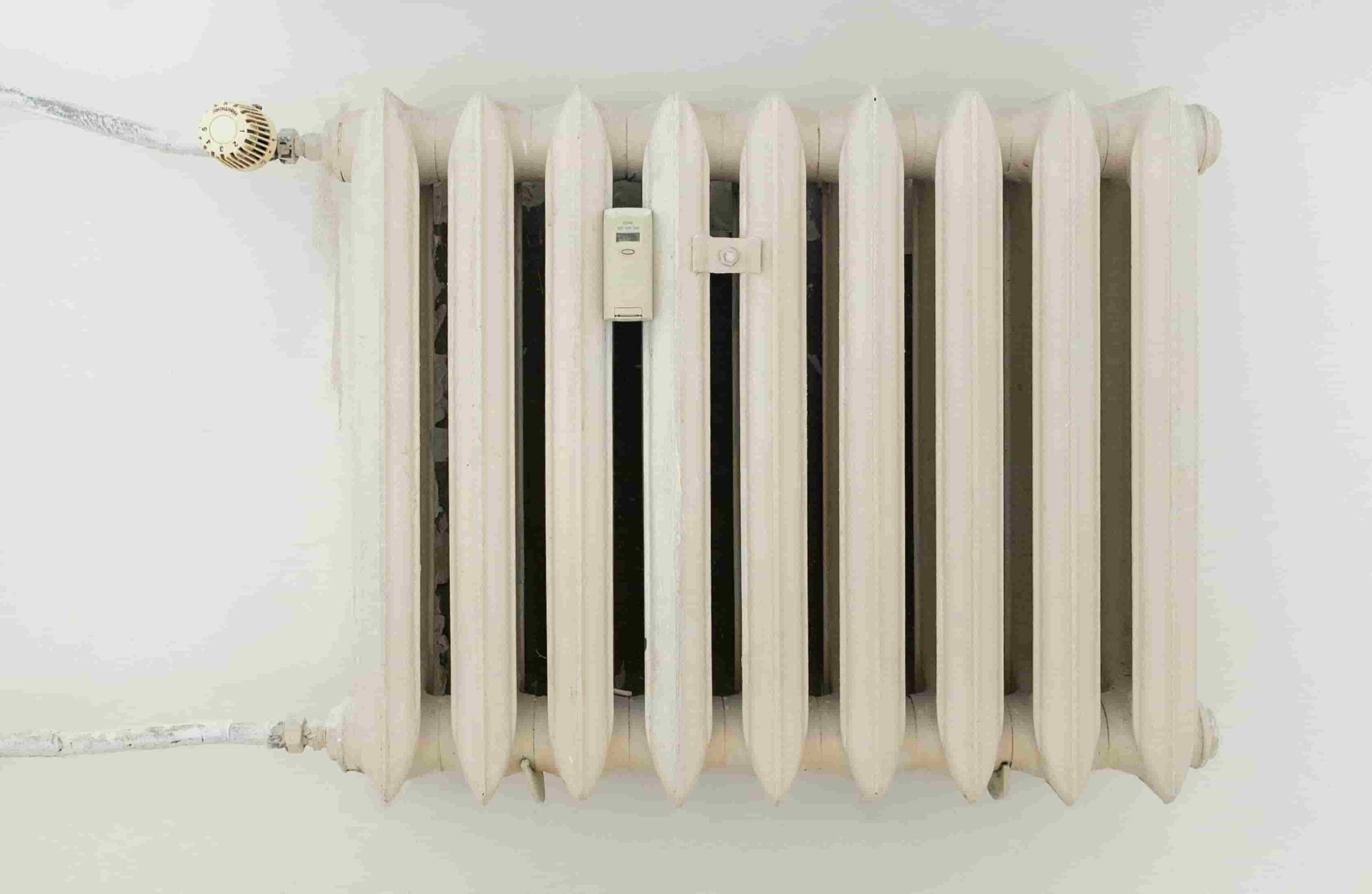 A white metal radiator with seven vertical sections, viewed from above and connected by pipes on both ends against a plain wall, is a subtle reminder of the cost of ignoring annual furnace tune ups