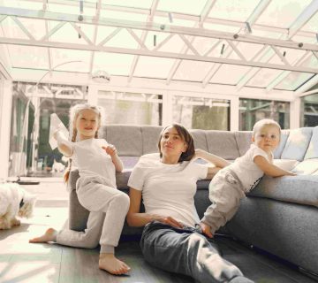 An adult and two young children relax on the floor next to a sofa in a bright, glass enclosed room with a small dog nearby, enjoying the comfort that comes from good indoor air quality in Irvine 