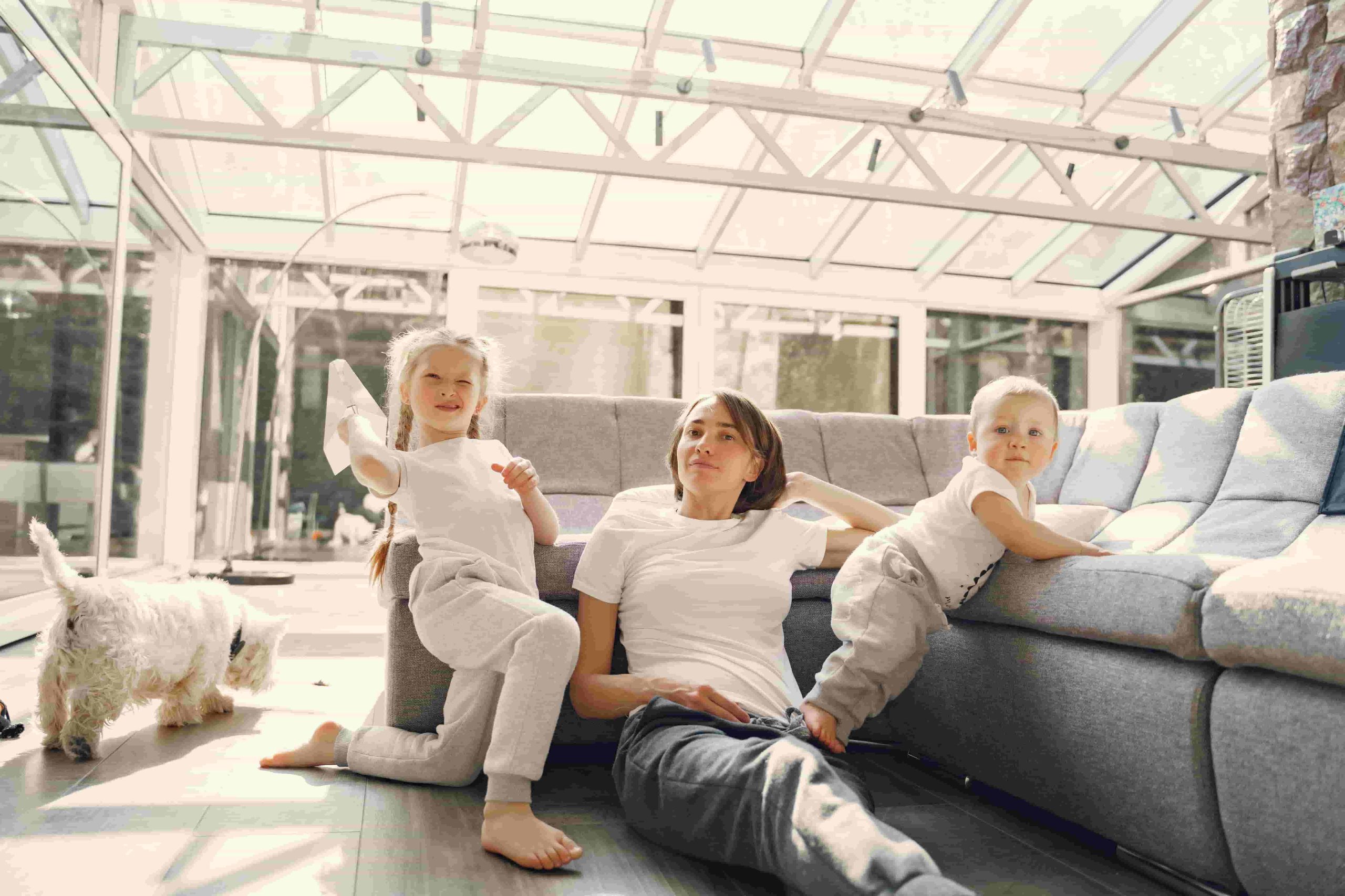 An adult and two young children relax on the floor next to a sofa in a bright, glass enclosed room with a small dog nearby, enjoying the comfort that comes from good indoor air quality in Irvine 