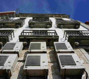 Several air conditioning units are mounted on the exterior wall of a building with multiple balconies, viewed from below against a clear blue sky—prompting the question: What are the signs of a refrigerant leak in my AC in Irvine?