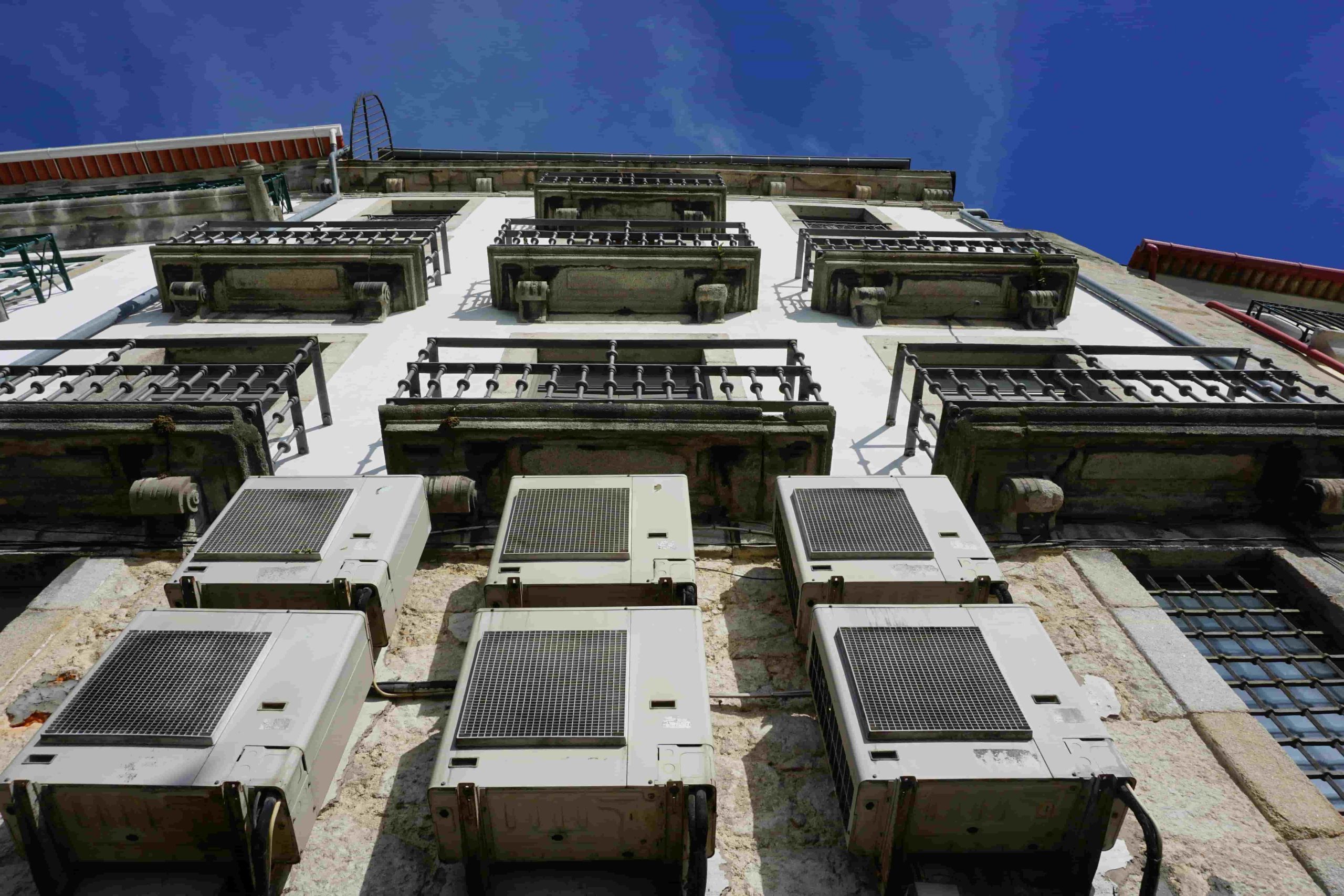 Several air conditioning units are mounted on the exterior wall of a building with multiple balconies, viewed from below against a clear blue sky—prompting the question: What are the signs of a refrigerant leak in my AC in Irvine?