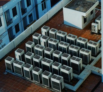 Rows of air conditioning units are installed on a tiled rooftop next to a small utility structure and a surrounding white wall 