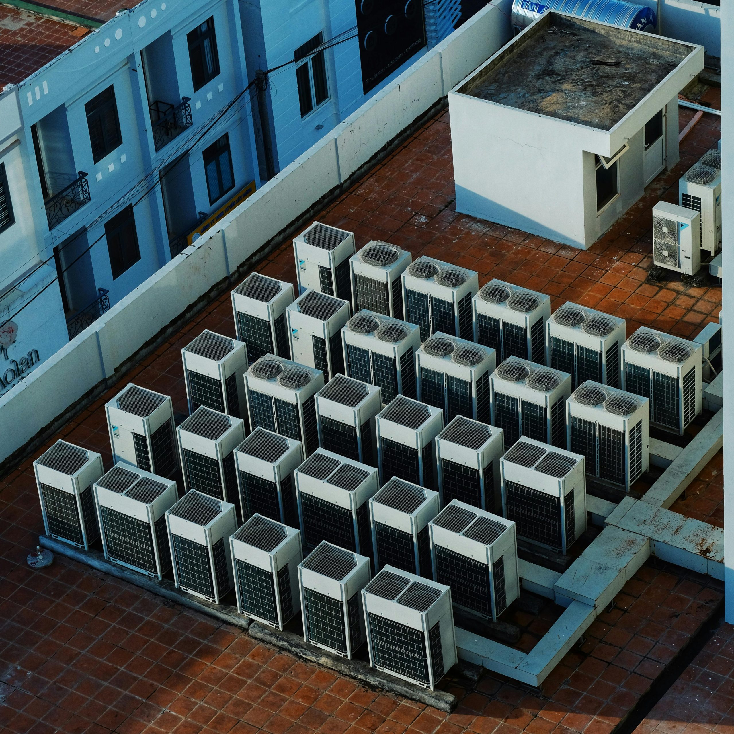 Rows of air conditioning units are installed on a tiled rooftop next to a small utility structure and a surrounding white wall 