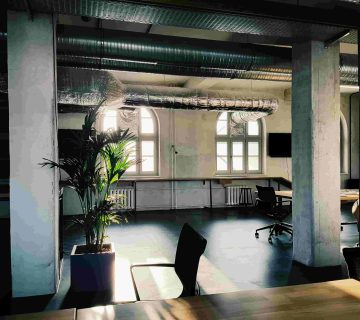 A modern, sunlit office space with large arched windows, exposed air ducts, potted plants, and empty desks and chairs 