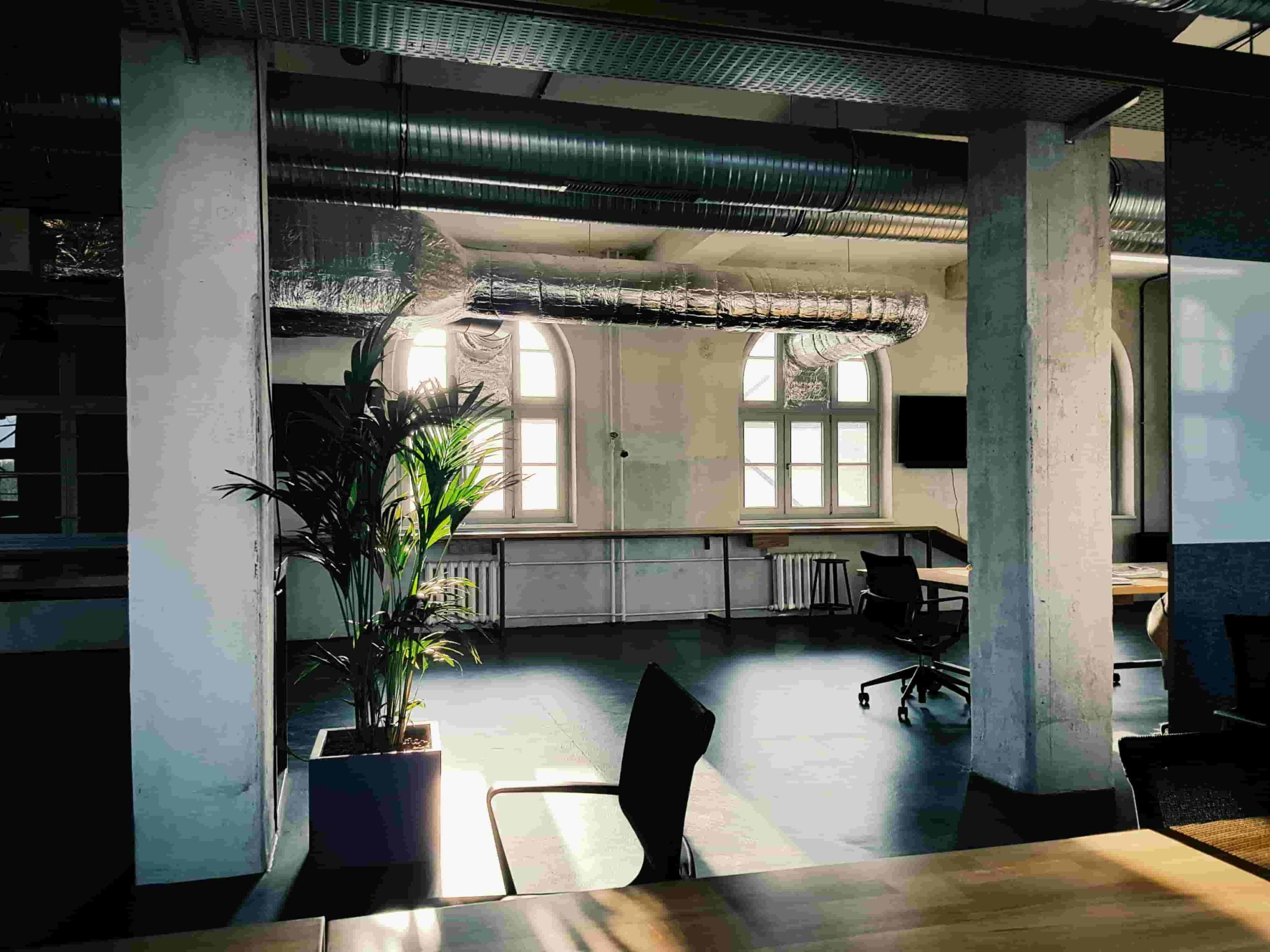 A modern, sunlit office space with large arched windows, exposed air ducts, potted plants, and empty desks and chairs 