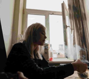 A person with long blonde hair sits at a table indoors, holding a cigarette with smoke rising near an open window with curtains, perhaps wondering: How can I improve air quality in my Irvine apartment? 