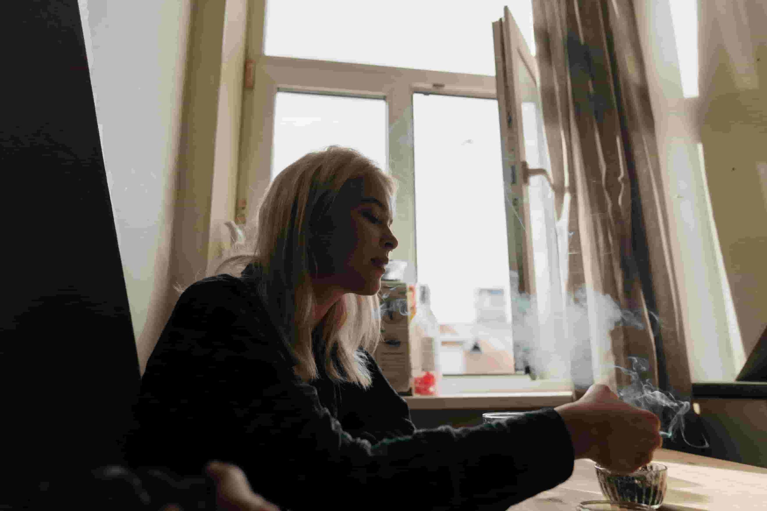 A person with long blonde hair sits at a table indoors, holding a cigarette with smoke rising near an open window with curtains, perhaps wondering: How can I improve air quality in my Irvine apartment? 