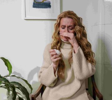 Woman with long hair sitting on a chair, wiping tears with a tissue, appearing upset or emotional next to a green plant—wondering, "How can I reduce allergens in my Irvine home? 