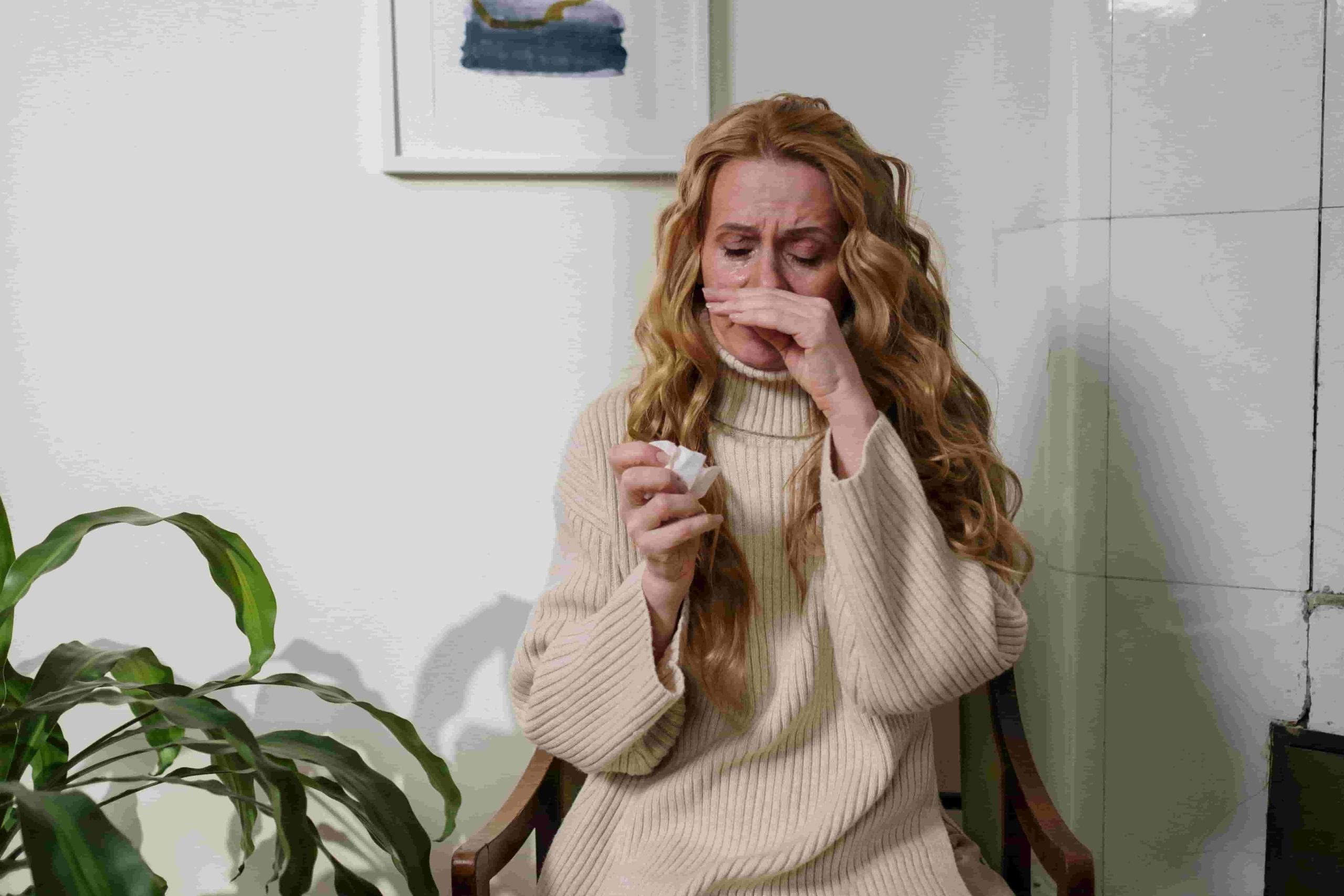 Woman with long hair sitting on a chair, wiping tears with a tissue, appearing upset or emotional next to a green plant—wondering, "How can I reduce allergens in my Irvine home? 