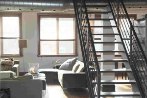Modern loft apartment interior with a metal staircase, large windows with blinds, two gray sofas, and a coffee table. Natural light fills the open living space—prompting the question: Is it worth repairing air ducts or should I replace them?.