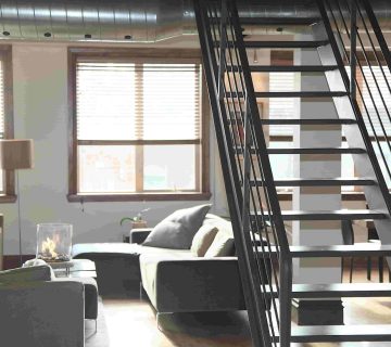 Modern loft apartment interior with a metal staircase, large windows with blinds, two gray sofas, and a coffee table  Natural light fills the open living space—prompting the question: Is it worth repairing air ducts or should I replace them? 