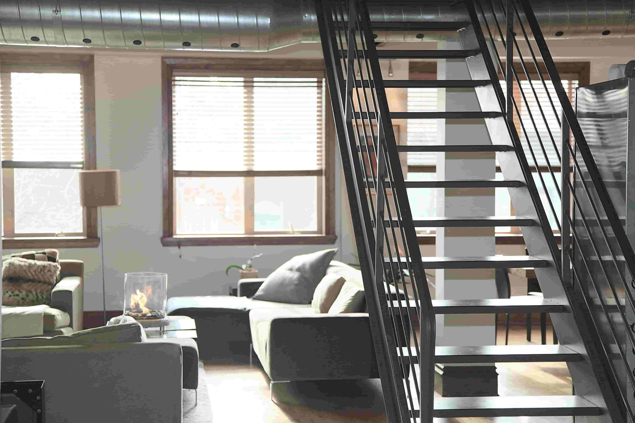 Modern loft apartment interior with a metal staircase, large windows with blinds, two gray sofas, and a coffee table  Natural light fills the open living space—prompting the question: Is it worth repairing air ducts or should I replace them? 