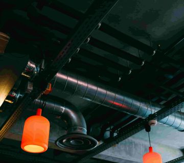 Exposed industrial ceiling with metal pipes and ductwork, illustrating what are the different types of air ducts, plus two orange hanging pendant lights 