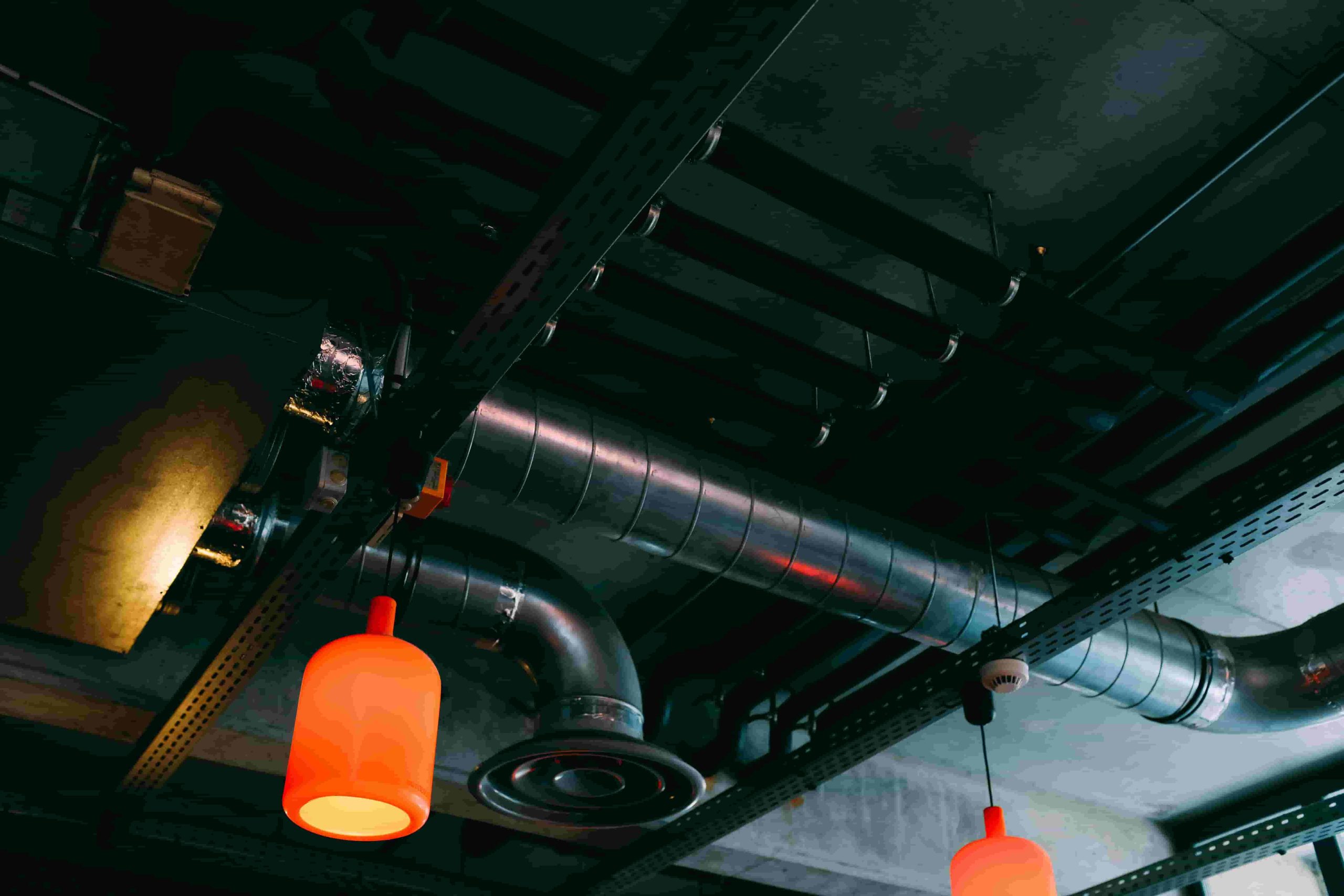 Exposed industrial ceiling with metal pipes and ductwork, illustrating what are the different types of air ducts, plus two orange hanging pendant lights 
